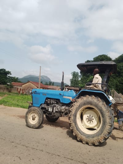 A blue tractor is parked on a paved road in Serti, Nigeria. The tractor has a man driving it, who is wearing a light-colored shirt. The tractor has large, rugged tires and a protective canopy over the driver's seat. In the background, there are several buildings with red-tiled roofs, some trees, and a mountain range in the distance. The sky is partly cloudy. The scene appears to be in a rural or semi-rural area.
