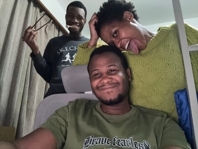 Three individuals are visible in an upside-down selfie. The first individual, wearing an olive-green t-shirt, is positioned at the top center. Below, a second person in a light green sweater is sticking out their tongue. To the right, the third individual is holding a mirror and wearing a black long-sleeved Skechers shirt. The background includes a gray office chair and a beige curtain. A blue bag and white frame is partially visible in the background. The location is unknown and the city of Daxing District, China, cannot be confirmed.