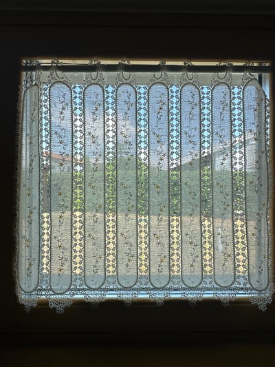 The image shows a close-up of a white lace curtain hanging in a window. The curtain has a scalloped bottom edge with intricate floral patterns. Vertical bands of lace with diamond-shaped cutouts run throughout the curtain, interspersed with embroidered floral sprigs. Through the semi-transparent fabric of the curtain, a view of green foliage and a house with a terracotta roof is visible. The scene appears to be set during the daytime, with natural light illuminating the interior through the window. The overall impression is one of a domestic interior with a view of an outdoor garden. The location of the image in Trieste, Italy, is not directly discernible from the visual elements alone, as the architecture and landscape are not distinctly identifiable as belonging to that specific city without external information.
