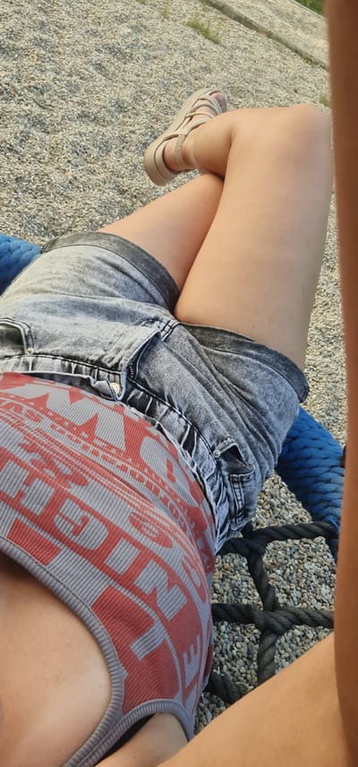 The image shows a close-up of a person's legs and torso, wearing denim shorts and a patterned tank top. They are sitting on a swing with thick ropes and a blue net, with a surface of small pebbles beneath. The lighting suggests it is daytime, possibly late afternoon given the warm tones. The scene appears to be outdoors, perhaps a park or playground, with gravel spread on the ground. The individual's legs are crossed, and their right foot is adorned with a light-colored sandal. The tank top has a repeating red pattern on a gray background. The image is shot from a high angle, looking down. No text is visible in the image. The overall impression is relaxed and casual, with a focus on summer attire and outdoor activity. The specific location context provided is Bratislava, Slovakia.