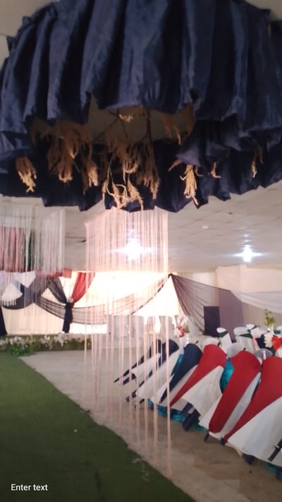 The image depicts an unoccupied indoor event hall in Gudi, Nigeria, prepared for a gathering. A large, dark blue ruffled fabric fixture with dangling light brown fibrous material hangs prominently from the ceiling in the upper foreground. The white ceiling above features multiple recessed light fixtures. Two large, illuminated chandelier-like structures, composed of numerous vertical, translucent strings, are suspended from the ceiling in the mid-ground.
The background shows walls draped with white, dark blue/black, and red fabrics, arranged in swags and folds, forming decorative backdrops and partitions. Additional white vertical string decorations are visible on the left side of the background.
Rows of unoccupied chairs, draped with white, red, and dark blue fabric covers, are arranged on a light beige floor surface towards the right side of the frame. Hints of teal fabric are visible beneath some chair covers. Partially visible tables display floral centerpieces featuring red and white flowers.
The floor in the immediate foreground consists of a strip of artificial green turf on the left, transitioning to the light beige solid floor surface that extends towards the background. A series of thin, light-colored vertical strands, possibly decorative strings, form a pathway or barrier in the mid-ground, extending towards the right. No individuals or human activity are depicted in the scene.