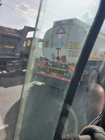 The image depicts an outdoor scene on an asphalt road in Kinanie ward, Kenya, viewed from inside a vehicle through a windshield. The primary subject is the rear of a large white tanker truck, stationary or moving slowly. The tanker features hazard placards, including a large red triangle, a red 'T' symbol, and a red diamond indicating flammable liquid (Class 3) with UN number 1203. Text on the tanker includes "ATTENTION," "EMERGENCY CALL" with Kenyan phone numbers (+254 722 798XXX, +254 723 068XXX), and "DANGER." The license plate "ZD 6892" is affixed to the rear bumper, along with reflective orange and red striped tape and an 80 km/h speed limit sign. To the left of the tanker, the partially visible rear of a black dump truck is present, loaded with dark, loose material, likely soil or gravel, and a yellow bucket hangs from its side. A portion of a white truck cab is discernible between the two main vehicles. The sky overhead is bright and partly cloudy, with visible power lines. To the far left, an elevated area of loose earth is visible. The immediate foreground shows streaks and reflections on the windshield, along with interior elements of the observer's vehicle and a partial reflection or view of a human arm/head on the right. No direct human actions are clearly visible outside the vehicles.