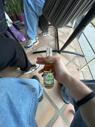 Captured from an overhead perspective, the image depicts a group of people sitting outdoors on a tiled terrace. In the foreground, a hand holds a partially consumed bottle of Somersby apple cider. The terrace is furnished with metal tables and chairs. Visible at the bottom left corner, a purple shopping bag rests beside a pair of sneakers. The location is Celorico de Basto, Portugal. The people are wearing casual clothing, including jeans and sneakers.
