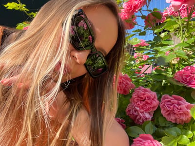 The image shows a close-up of a young woman with long blonde hair, wearing sunglasses. The sunglasses reflect pink roses and green foliage. She is outdoors, with a rose bush prominently featured in the foreground and background. The sky is visible and appears blue. The overall impression is of a sunny day. The location is identified as Novopokrovskaya, Russia.