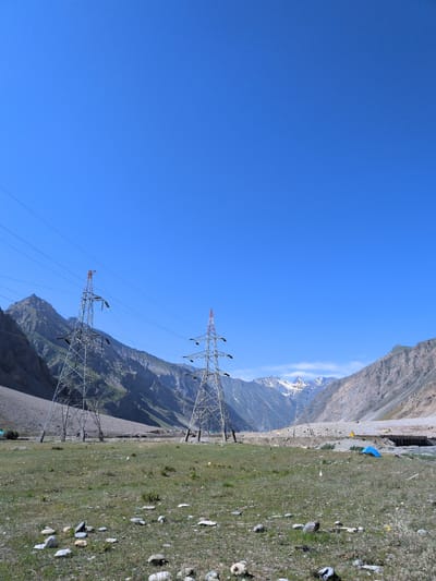 This is a landscape photograph taken in Matayeen, India, under a clear blue sky. The foreground features a grassy field scattered with rocks. Two large, latticed metal electricity pylons stand prominently in the mid-ground, with power lines extending between them and into the distance. Beyond the pylons, a valley unfolds with steep, rocky mountains on either side, some of which are snow-capped in the distance. The terrain is rugged and sparsely vegetated. In the far mid-ground, a blue tent is visible on the right side of the frame, near what appears to be a body of water or a riverbed and a bridge. The overall impression is one of a remote, mountainous region with signs of infrastructure.