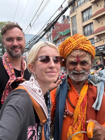 A selfie shows three people in an urban environment. A gray-bearded man wearing a yellow patterned turban and orange robes is at the top of the frame. He has painted markings on his face. Beneath him is a blonde woman with sunglasses, a nose ring, and a patterned scarf. At the bottom left is a man with a beard and a mark on his forehead. Buildings and overhead wires are visible in the background. A sign for "MAHARAJA THE ROYAL CASINO" is also visible. The location is Pokhara, Nepal.