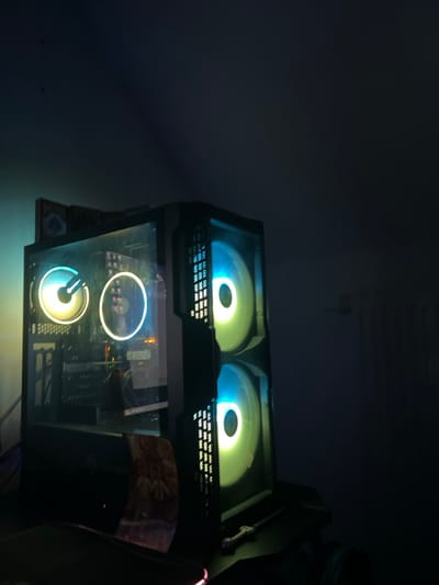 The scene, located in the city of Benningen, Germany, displays a dimly lit interior focused on a desktop computer tower. The computer case is black, featuring a transparent tempered glass side panel and a perforated front panel. Four circular RGB fans are visible: two internal fans with yellow/green and blue illumination, and two front-mounted fans, also glowing yellow/green and blue, visible through the perforated panel. A blue box, partially identifiable as a "Bicycle" brand playing card deck, rests on top of the computer case. The computer is situated on a dark desk surface. To the immediate right of the computer, a small, silver-hilted object resembling a miniature sword or letter opener is visible on the desk. A segment of a mousepad with a red illuminated edge is present in the lower-left foreground. The background consists of a dark wall, with the faint outline of a radiator or similar fixture visible on the right side. No individuals are present.