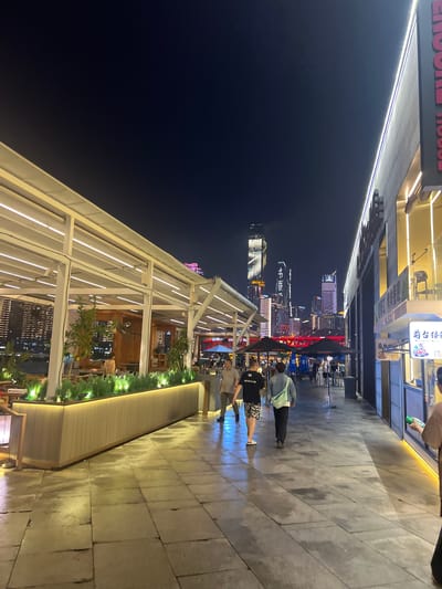 This media file captures a nighttime scene on a city street in Jiangbeicheng Subdistrict, China. The perspective is angled, with the foreground dominated by a modern, covered walkway or patio area with white structural beams and integrated LED lighting. This area appears to be an outdoor seating section of a restaurant or establishment, with plants and seating visible within.
Beyond this structure, the scene opens up to a bustling city street at night. The background features a vibrant cityscape with numerous illuminated skyscrapers and buildings, indicative of a well-developed urban environment. A red bridge is visible spanning across a body of water or a large thoroughfare, further enhancing the urban landscape.
Several people are present in the scene, walking along the paved sidewalk or seated in the outdoor area. They are casually dressed, suggesting a relaxed evening outing. The ground appears wet, possibly from recent rain, reflecting the ambient light and adding to the nighttime atmosphere.
Notable details include the bright artificial lighting from buildings and signage, typical of a city at night. The presence of illuminated skyscrapers suggests a significant urban center. The overall mood is lively and energetic, with the city lights creating a dynamic backdrop.
There is some visible text in the image. Above the cityscape, on signs attached to buildings, there are Chinese characters. On a sign closer to the foreground, also with Chinese characters, there appears to be a logo or establishment name. One sign, seen partially cropped in a hint image, seems to read "ENCORE HOUSE" in English.