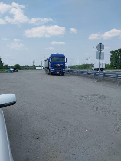 In the city of Troitskoe, Russia, a blue MAN truck with a trailer is parked on a gravel lot. The truck is facing right, with its front visible. The lot is surrounded by trees and a blue sky with white clouds. In the background, there is a white box truck and several other vehicles. A white car's side mirror is visible in the foreground. The scene appears to be a roadside or a parking area.
