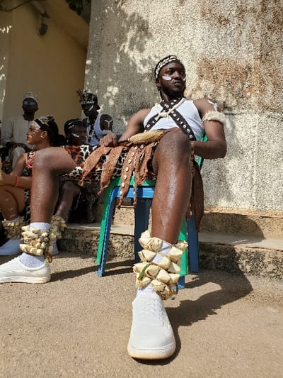 The scene depicts an outdoor gathering in Bar Gada, Nigeria, featuring multiple individuals in cultural or ceremonial attire under bright sunlight. In the foreground, an adult male with dark skin is seated on a light blue plastic stool. He wears a white tank top, a dark strap chest harness with white cowrie shells, and a skirt-like garment comprising animal print fabric layered with dark, dried materials, also adorned with cowrie shells. White ritualistic paint marks are present on his face, and he wears a cowrie shell headband and a furry white armband on his right bicep. His skin is visibly oiled or glossy. He wears white socks and white sneakers, with distinct ankle adornments made of light-colored, pyramidal elements fastened with green string over the socks. He gazes towards the upper right. To his left, partially obscured, an adult female is seated, wearing an animal print garment, a cowrie shell headband, and white face paint, with similar footwear and ankle adornments. In the background on the left, at least three additional individuals are seated. One man wears white traditional clothing and sunglasses, while another wears a white sleeveless top, a cowrie shell headband, and white face paint. The ground is a light brown, compacted earth or concrete surface. The background consists of a textured, light-colored concrete or stucco wall with visible weathering and dark stains, and a lighter structure with an overhang on the far left. Distinct shadows from the figures and overhead foliage are cast on the ground and walls.