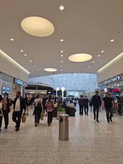 The image depicts the interior of the Weserpark shopping mall in Bremen, Germany. Several people are walking through the mall, some are holding shopping bags, and others are eating ice cream. The mall features a modern design with a high, illuminated ceiling and various storefronts. The floor is tiled with a speckled pattern. The lighting is bright and even, typical of a commercial indoor space. In the foreground, a metal trash receptacle is visible. The overall atmosphere appears to be a busy, everyday scene within a large shopping center.