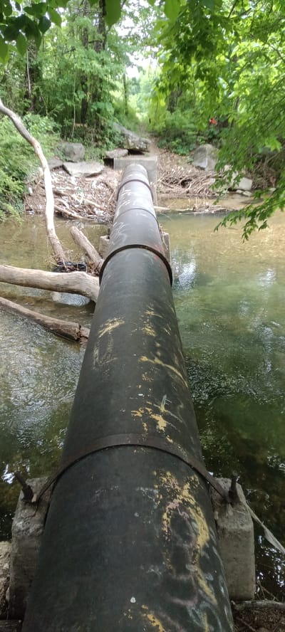 A metal pipe spans a shallow stream in a wooded area. The pipe is supported by concrete blocks at intervals. The stream is clear with visible rocks and debris. The surrounding environment is lush with trees and foliage. A path or trail is visible leading away from the stream. The location is in Tahlequah, United States of America.