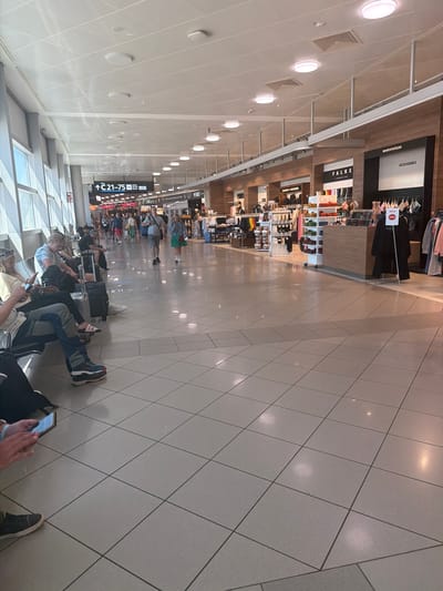 This is a rotated image taken inside an airport terminal at Schwechat, Austria. The scene shows a long corridor with shops on one side and a waiting area with people sitting on the other. The floor is tiled with a light-colored, possibly ceramic or stone material, laid in a diamond pattern.
Several people are visible, most of whom are seated in rows, facing away from the shops and towards the viewer. They are dressed casually, with luggage placed around them. Some are looking at their phones, while others appear to be waiting patiently. A few individuals are standing or walking in the distance, near the shops.
The shops visible include brands like "FALKE," "TOMMY HILFIGER," and what appears to be a duty-free store. Signage within the shops indicates "ACCESSORIES" and "SALE" with a 50% discount. Further down the corridor, a directional sign reads "C 21-75" with an arrow, suggesting gate information. The ceiling is white and features circular light fixtures. The overall lighting suggests it is daytime, with artificial lighting supplementing natural light that might be entering from unseen windows. The atmosphere appears calm and typical of an airport waiting area.