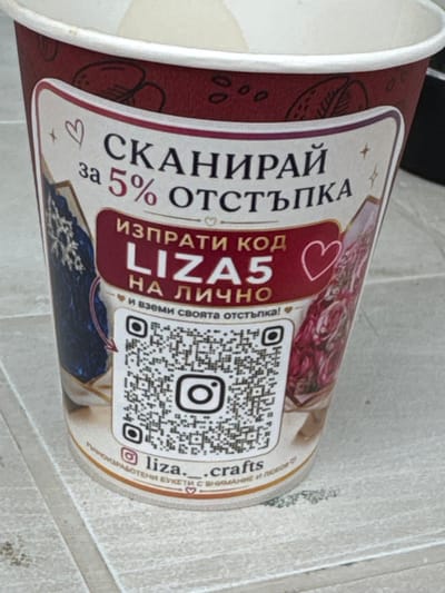 The image shows a close-up of a paper cup with a promotional design. The cup is primarily dark red with a cream-colored rim. The design features text and graphics, including a QR code and social media icons. The text, in Cyrillic script, appears to be an advertisement for a discount. Specifically, it reads "СКАНИРАЙ за 5% ОТСТЪПКА" which translates to "SCAN FOR 5% DISCOUNT". Below this, it says "ИЗПРАТИ КОД LIZA5 НА ЛИЧНО" meaning "SEND CODE LIZA5 PRIVATELY". There are also smaller text elements, including an Instagram logo and the handle "liza.__crafts", and a phrase "РЪЧНОРАБОТЕНИ БУКЕТИ С ВНИМАНИЕ И ЛЮБОВ" which translates to "HANDMADE BOUQUETS WITH CARE AND LOVE". The background is a light-colored, textured surface that appears to be a table or countertop. There are no people or animals visible in the image. The location is not identifiable from the provided image.