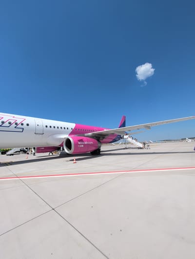 The image depicts a Wizz Air airplane parked on the tarmac at Frankfurt-Hahn Airport (HHN) in Lautzenhausen, Germany, under a clear blue sky with a single white cloud. The left side of the aircraft, featuring the airline's signature pink and white livery with a stylized "wizz" logo, is prominently visible. A large engine with "wizzair.com" written on it is also clearly visible on the right side of the aircraft. A set of mobile stairs is connected to the plane's rear door, with several passengers ascending or descending. Other ground vehicles and more aircraft are visible in the background, suggesting a typical airport environment. The ground is concrete, marked with yellow and red lines.