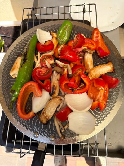 The image shows a close-up, overhead view of a grill basket filled with various vegetables and mushrooms, likely prepared for grilling. The subjects are predominantly the food items: sliced red bell peppers, green chili peppers, halved cherry tomatoes, thick slices of white onion, and several types of mushrooms, including what appear to be king oyster mushrooms and enoki mushrooms. The vegetables are cut into bite-sized pieces, and some show signs of charring, indicating they have been on the grill.
The grill basket itself is a round, metal mesh with a patterned surface and a black handle. It is placed on top of a metal grill rack, which is part of a larger outdoor grill. In the background, a white plate is partially visible on the left, and a blurred background suggests an outdoor setting, possibly a patio or backyard. The lighting suggests it is daytime, with natural light illuminating the food. There are no people visible in the frame.
The activity taking place is the preparation of food for grilling, or the food is in the process of being grilled. The scene is set outdoors in a domestic environment. Notable details include the variety of colorful vegetables, the texture of the grilled mushrooms, and the organized arrangement of the ingredients within the grill basket. The location cues are general, indicating an outdoor cooking scenario. There is no visible text in the image. The overall impression is of healthy, appetizing food ready for a meal.