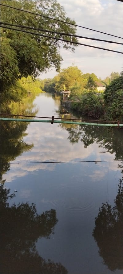 The image depicts a tranquil river flowing through a verdant landscape, likely in Mataram, Indonesia. The river's surface reflects the sky, clouds, and surrounding trees, creating a mirror-like effect. A small wooden structure with a yellow roof is visible on the riverbank, accompanied by lush vegetation. Overhead, several dark electrical wires traverse the frame, partially obscuring the sky. The overall atmosphere is serene and natural, with the golden hour light casting a warm glow on the scene.