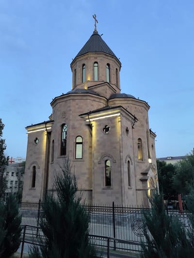 An external, low-angle, full-shot photograph captures a stone church with a dark, conical dome topped by a cross. The church's facade is illuminated by several exterior lights. The sky is clear and a pale blue, indicating either dawn or dusk. The church is surrounded by a black metal fence, in front of which are several evergreen trees. In the background, the upper floors of a multi-story building are visible, with trees and other structures interspersed. The image appears to be taken in the city of Veshenskaya, Russia.