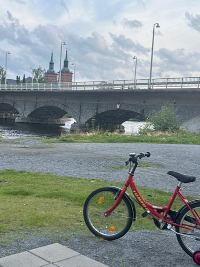The image depicts a red child's bicycle parked on a gravel path next to a grassy area. In the background, a stone bridge with multiple arches spans a body of water. Beyond the bridge, two church towers with green spires are visible against a cloudy sky. The overall scene appears to be an outdoor public space in Vammala, Finland, likely near a river or canal.