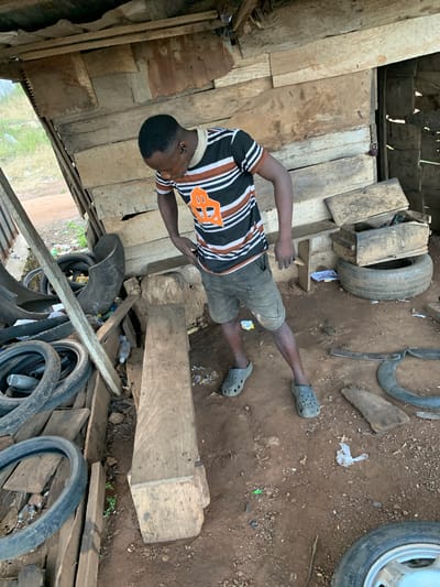 This media file shows a person in an outdoor, likely workshop or storage area, with a dirt ground and wooden structures. The subject, a young person wearing a black, white, and orange striped t-shirt and dark shorts with grey Crocs, is in a supine position, with their head facing right and their legs extended towards the left. Their right arm is bent, resting on their chest, while their left arm is extended downwards.
The scene is cluttered with various objects, including several tires of different sizes, some of which appear to be old or discarded. Wooden planks and debris are scattered around the area, contributing to a rustic and somewhat unkempt environment. The lighting suggests it is daytime, with natural light illuminating the scene. There are no clear indications of weather conditions, but the dirt ground and general appearance suggest a dry, possibly dusty environment.
The overall impression is of a utilitarian space, possibly a repair shop or a place where salvaged materials are stored. The subject's pose could suggest rest or contemplation. No text is visible in the image. The location context is Kabba, Nigeria.