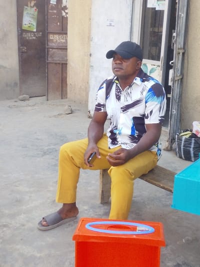 A man in a black baseball cap, patterned shirt, and yellow pants sits on a wooden bench. He wears gray sandals and holds a phone in his left hand. Beside him are orange and blue plastic bins. Behind him is a weathered building with two doors, one with a "CLOSE GATE AT 11:30 PM" sign. A black and white striped bag is also visible. The man appears to be looking off to the right.