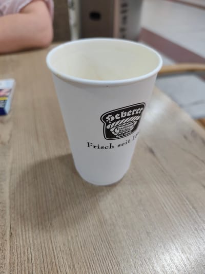 This media file features a close-up, slightly angled shot of a white paper coffee cup with a black logo and text. The cup is positioned on a light-colored wooden table with a distinct wood grain pattern. In the blurred background, a person's arm and shoulder, wearing a light pink garment, are visible on the left side of the frame. To the left of the cup, a pack of cigarettes, possibly with red and blue branding, is partially visible. The setting appears to be indoors, on a table, suggesting a cafe or a similar establishment. The lighting is soft and diffused, indicating it could be daytime, but no specific time can be determined. The overall mood is casual and relaxed.
The logo on the cup is in black and appears to be a stylized depiction of a braided rope or bread, enclosed in an oval shape. Within the logo, there is text that reads "Geberer Traditions Bäcker" and "Seit 1891". Below the logo, the text "Frisch seit 1891" is visible, also in black. The cup itself is clean and empty. The presence of the coffee cup, cigarettes, and a person's arm suggests a break or a casual moment. Given the text on the cup, which includes German words and a historical date, the location is likely Germany, specifically Frankfurt as indicated in the location context.