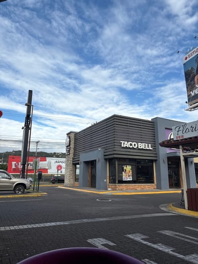 The scene shows a Taco Bell restaurant in Pozos, Costa Rica, located at 9.952 latitude and -84.194 longitude. The building is a modern structure with a gray exterior and brown accents. The Taco Bell sign is prominent and visible on the front of the building. There is a parking lot in front of the restaurant, and a few cars are parked. The sky is blue and partly cloudy. There are some power lines overhead. The environment is urban and the scene is empty.