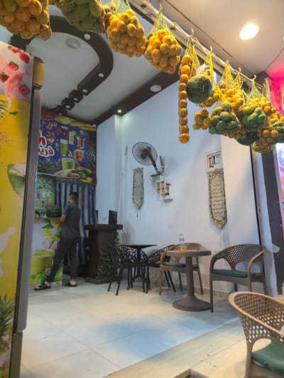 The image depicts the interior of a juice bar or similar establishment in Al Hadaba, Egypt. The scene is brightly lit, with a white tiled floor and light blue walls. Hanging from the ceiling, suspended in yellow netting, are large quantities of oranges and other round fruits, some green and some yellow. A man, wearing a grey t-shirt and dark trousers with sandals and white socks, is standing to the left, facing away from the camera. He appears to be interacting with a display or counter. Behind him, a large mural adorns the wall, showcasing various fruit drinks and ingredients like strawberries, pineapples, and oranges.
In the center of the room, a dark wooden counter or display unit is visible, with greenery cascading down its sides. Nearby, two tables are set up for seating. One is a small, round, brown table with a single bottle of water on it. Surrounding the tables are several chairs made of woven material with green cushions. A black oscillating fan is mounted on the wall to the right of the entrance. Two decorative tapestries with Arabic script hang on the wall, one on either side of a small white shelving unit with a few decorative items. A security camera is mounted high on the wall above the mural. The overall impression is of a clean and inviting establishment.