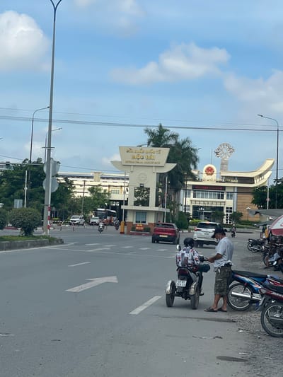 This is an image of the Moc Bai International Border Gate in Ben Cau, Vietnam. The image depicts a street scene with vehicles and people. A large, modern building with a prominent archway and signage marks the border crossing. The signage on the archway is in Vietnamese and English, indicating "CUA KHAU QUOC TE MOC BAI" and "MOC BAI INTERNATIONAL BORDER GATE". The building behind the archway has a central section with a Vietnamese flag and more signage that appears to be welcoming visitors.

In the foreground, a person is standing next to a three-wheeled motorcycle, interacting with another person seated on the motorcycle. The person standing is wearing camouflage shorts and a light-colored checkered shirt. The person on the motorcycle is wearing a patterned shirt and a helmet. Several other motorcycles are parked on the right side of the image.

The street is paved with asphalt and has white lane markings, including an arrow indicating a direction. Traffic appears to be moving through the border gate, with cars and motorcycles visible. Lush green trees and palm trees are present around the border gate area, suggesting a tropical climate. The sky is partly cloudy, with white clouds against a blue background. The overall scene suggests a busy and active border crossing point.