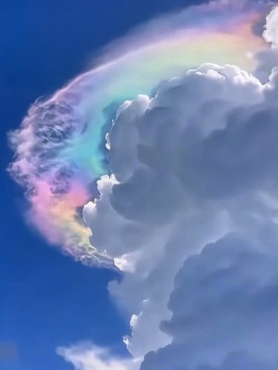 The image captures a sky with a natural phenomenon, possibly a circumhorizontal arc or cloud iridescence, appearing as a band of soft, pastel rainbow colors. This colorful display is set against a backdrop of a clear blue sky on the left, transitioning to dense, puffy white and gray clouds occupying the right side of the frame. The clouds are illuminated from above, casting shadows that create a sense of depth and volume. The vibrant colors are concentrated along the upper edge of the clouds, suggesting the interaction of sunlight with ice crystals or water droplets within the atmosphere. The image does not show any specific location within Riverhead, United States of America, nor does it depict any people, objects, or actions.