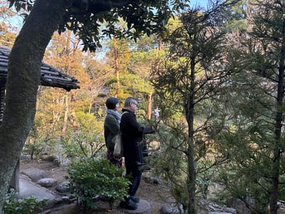 Two adults, a man and a woman, stand in a densely wooded area in Minato City, Japan (latitude: 35.662, longitude: 139.717). The woman stands to the left of the man and is wearing a dark green jacket, a white shirt, and a small black bag hanging from her shoulder. She appears to be holding something small in her hand. The man, who is taller and standing on the right, is wearing a dark blue jacket and is holding his left hand on his hip. He appears to be looking out towards a small, indistinct figure in the distance. The pair are standing on a rocky ground slope covered in green bushes and trees. The image is taken from a low angle, with the camera focused on the two adults. In the background, one can see a small, wooden hut, tree trunks and foliage. The image is shot with a telephoto lens, with the foreground slightly blurred.