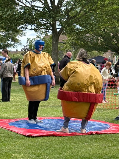 Two individuals are participating in a sumo wrestling activity on a circular inflatable mat. The mat is red on the outer edge and blue in the center, with a white swirl pattern. The participants are wearing inflatable sumo wrestling costumes that are yellow, with blue padding around the waist and red padding around the lower part of the costume. The person on the left is facing forward and wearing a blue helmet. The person on the right is facing away from the camera. Both individuals are standing on the mat. In the background, there are trees, buildings, and other people observing. The ground is covered in green grass. The scene appears to be an outdoor event, possibly a festival or fair, set in a park or open field. The location is identified as Great Yarmouth, United Kingdom.