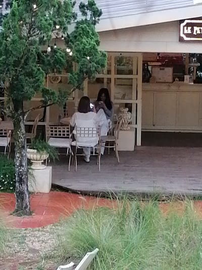 This image depicts an outdoor seating area of a cafe, likely located in Lang Biang - Đà Lạt, Vietnam, given the presence of decorative foliage and possibly the architectural style. Two women are seated at tables. The woman in the foreground, viewed from behind, is wearing a white top and pants and is seated in a chair with a patterned back. She appears to be looking at her phone. The woman behind her, to her left and further away, is wearing a blue top and is also seated. She is also looking down, possibly at her phone or another object. In the background, the cafe structure is visible, with a white exterior and large windows or glass doors. A lit sign with white lettering on a dark background is mounted on the upper right of the building, possibly indicating the cafe's name. The foreground shows a red tiled area with a tree and a decorative planter. Lush green grass and foliage are visible in the lower part of the image. The overall impression is that of a relaxed outdoor cafe setting.