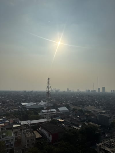 This is an aerial, high-angle, wide shot of a densely populated urban environment in South Jakarta, Indonesia, under a hazy, overcast sky. The sun, obscured by atmospheric conditions, shines brightly in the upper center of the frame, casting a starburst effect. In the foreground, various low-rise buildings with tiled roofs, some appearing residential and others possibly commercial or industrial, are visible. A prominent telecommunications tower stands out to the right of center, its metallic structure reaching high into the sky. In the mid-ground and background, a sprawling cityscape unfolds, characterized by a mix of residential areas, larger buildings, and other tall structures, all softened by the pervasive haze. The overall impression is one of a vast, densely packed urban landscape viewed during daylight hours, with the sun's presence creating a lens flare effect. No individuals or explicit actions are discernible.