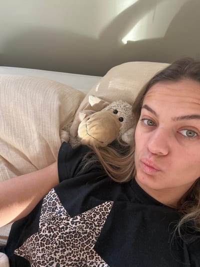 An adult female, characterized by light brown hair and light-colored eyes, is depicted in a close-up selfie. She lies on light-colored, textured bedding, wearing a black short-sleeved t-shirt with a large leopard-print star design on the front. Her lips are puckered, and she looks directly at the camera. To her left, resting on the bedding, is a light brown stuffed sheep toy with black button eyes. The scene is illuminated by natural light entering from the left, casting distinct shadows on a plain, light-colored wall in the upper background. The location is Veauchette, France.