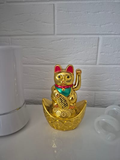 This is a close-up, eye-level, indoor photograph of a golden Maneki-neko (beckoning cat) figurine placed on a white, reflective surface. The figurine is positioned in the center of the frame, sitting on a golden boat-shaped object adorned with coin imprints. The cat has its right paw raised in a beckoning gesture and holds a golden ingot in its left. It wears a green bib and a red collar with a small golden bell. The background consists of a white brick-patterned wall. To the left of the figurine, a portion of a white cylindrical object, possibly a lamp or humidifier, is visible. To the right, two translucent white objects, appearing to be coiled tubes or rings, are present. The lighting is bright and casts soft shadows. The location of this scene is not specified as Barbing, Germany, or any other city.