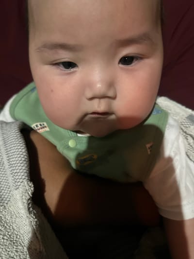 The image shows a close-up of a baby's face, tilted to the side. The baby has fair skin and dark eyes. They are wearing a light green onesie with embroidered patches. A white lace-trimmed garment is visible around the baby's shoulders. The background is a dark, blurred surface, possibly a blanket or bedding, with a darker area suggesting a shadow or a person holding the baby. The lighting is dim, indicating an indoor setting, possibly at night or in a dimly lit room. There is no discernible text in the image. The baby appears calm and relaxed. Given the location context of Тайгараев, Kyrgyzstan, the scene likely depicts a domestic setting within that region.
