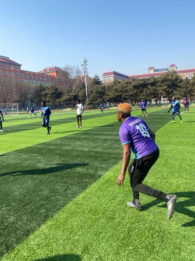 The image captures an outdoor football (soccer) field at Liaoning University of Technology in Taihe District, Jinzhou, China, under a clear, bright blue sky. Approximately ten male individuals are distributed across the artificial green turf field, engaged in what appears to be a game or training session. In the immediate foreground, an individual with blonde/light orange hair, wearing a purple and light blue jersey with "SHADOW" and the number "88" on the back, black athletic pants, and dark cleated shoes, is observed from behind in a crouched, ready stance, facing towards the left side of the frame. Other players are visible in the mid-ground and background; one in a blue and black striped jersey with "29" is walking towards the right, another in a white t-shirt and black shorts stands centrally, and several others in blue or purple jerseys are positioned across the field. A white football goal post is discernible in the far-left background. Beyond the field, a line of evergreen trees and multi-story academic buildings with reddish-orange brick facades are visible, along with a tall telecommunications tower. Shadows are cast distinctly on the turf, indicating direct sunlight.