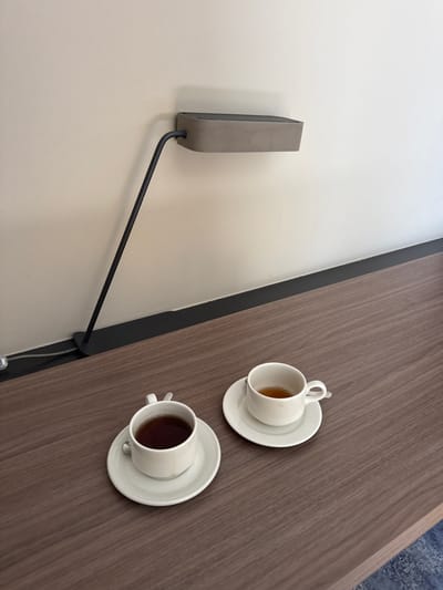 The image shows a wooden desk with two white teacups and saucers, each containing tea. A modern black lamp with a gray rectangular shade is attached to the wall behind the desk. The scene is lit by the lamp. The desk is positioned against a wall painted a light beige. A dark gray baseboard runs along the bottom of the wall. The flooring beneath the desk is not fully visible but appears to be carpet with a blue pattern. The setting is likely an indoor space, such as an office or a room in a residence. There are no people or animals present in the image. There is no specific information in the image to indicate its location in Ljubljana, Slovenia.