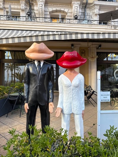 Two humanoid sculptures stand prominently in front of an ornate building in Nice, France. The sculpture on the left depicts a male figure wearing a glossy black suit and tie, with its head replaced by an oversized, smooth, light peach-toned pair of lips. The sculpture on the right portrays a female figure in a textured white V-neck top and matching trousers, with its head replaced by an oversized, glossy bright red pair of lips; two small white circular elements are visible on the upper red lip. Both sculptures feature light-toned hands.
In the immediate foreground, a low hedge of green shrubbery partially obscures the lower sections of the sculptures. Behind them, a paved patio area is visible with dark metal chairs, some stacked or partially arrayed, suggesting an outdoor seating establishment. The background features a multi-story, classically styled building with a light-colored facade, ornate architectural details including decorative pilasters, cornices, and balconies with black railings. A grey and white striped awning extends over the ground-level patio. Large glass windows and doors reflect palm trees and reveal an interior with a chandelier. A monogram, possibly "RA" or "AR," is visible above the main entrance. To the right of the sculptures, a white sign states "SCULPTEUR RICHARD MAS" and a phone number "06 72 65 33 46."