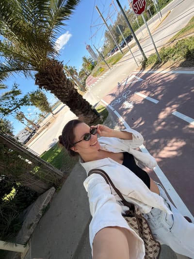 A selfie of a young woman wearing sunglasses, a white shirt, black top, and denim shorts stands next to a palm tree on a sunny day in Glifada, Greece. A street and a bike path with a stop sign are visible in the background. A construction crane can be seen in the distance.