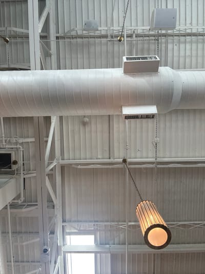 The image displays an upward view of an exposed industrial-style ceiling within an indoor commercial or public space in Highland Park, United States. The scene is dominated by white, encompassing the corrugated metal ceiling panels, structural steel beams, trusses, pipes, and electrical conduits.
A large, cylindrical white HVAC duct runs horizontally across the upper-middle section of the frame. Two smaller, rectangular white ventilation registers, each with a dark grille, are attached to the underside of this main duct. Two gray, box-shaped loudspeakers are mounted on the ceiling's support structures, one in the upper left and another in the upper right. A circular white device, consistent with a smoke detector, is affixed to a corrugated ceiling panel in the mid-left.
A prominent pendant light fixture hangs in the lower right quadrant, suspended by a dark metal chain. This fixture is cylindrical, featuring vertical ribbed translucent panels and dark metallic trim, and emits a warm yellow light from its open circular bottom. Other smaller, brass-colored fixtures with attached chains are visible higher up on the ceiling structure. Towards the bottom left, a rectangular opening reveals a bright, undifferentiated light source, indicating a window or skylight. No individuals or specific branding are present.