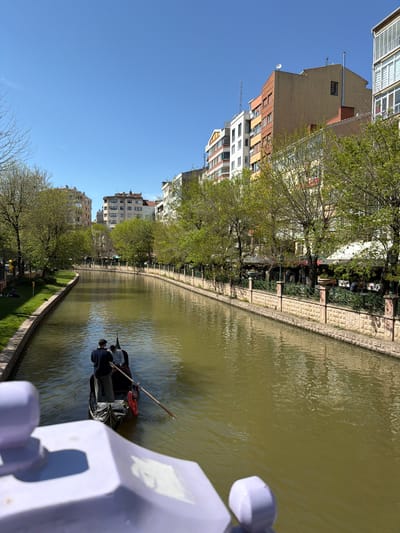 The image captures an urban canal scene in Eskisehir, Turkey, under a clear, bright blue sky. The canal, filled with green-brown water, stretches from the foreground into the distance.
In the middle of the canal, a dark-colored boat, resembling a gondola, is being actively poled by one person, while another person is seated as a passenger. A red life preserver or buoy is visible inside the boat. Ripples on the water surface reflect the daylight.
The right bank of the canal features a stone retaining wall topped with an ornate green fence. Behind this fence is a green park area lined with trees, some displaying fresh spring foliage. Interspersed among the trees are outdoor seating arrangements, including tables and chairs, suggesting cafes or restaurants alongside the canal.
On the left side of the canal, a series of multi-story buildings stand, showcasing varying architectural styles and colors, including beige, red brick, and white facades. Some buildings feature balconies, and several rooftops are adorned with satellite dishes and antennas.
The photograph appears to be taken from an elevated position, likely a bridge, with a light purple railing partially visible in the immediate foreground. The clear blue sky and strong light indicate daytime, likely morning or afternoon, with sunny weather. The overall scene depicts a calm and recreational urban environment. No discernible text is visible within the image.