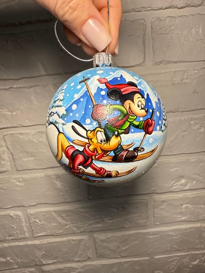 This is a medium shot, eye-level photograph of a hand holding a Christmas ornament. The ornament is round and painted with a winter scene featuring Disney characters Mickey Mouse and Pluto skiing. Mickey Mouse is in the foreground, wearing a red hat and green scarf, and holding ski poles. Pluto is behind him, also on skis. The background depicts snowy mountains and trees. The hand holding the ornament has light-colored fingernails. The ornament is suspended against a grey brick wall. There is no information provided to determine the location in Lipetsk, Russia.