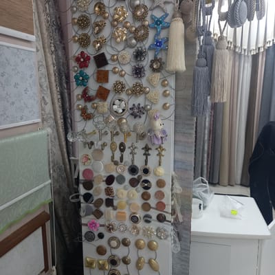 In a retail setting, likely a fabric or home decor store, a display of ornate curtain tiebacks and decorative buttons is presented against a backdrop of rolled fabric bolts. The tiebacks, featuring tassels in various colors and materials such as cream, grey, and beige, hang from a vertical metal grid. Below the tiebacks, a collection of decorative buttons and embellishments are displayed, including a small bear figurine. The items vary in material and design, with many featuring metallic finishes, rhinestones, and intricate patterns like flowers and butterflies. The overall impression is one of a well-organized retail display showcasing decorative accessories for home interiors. The location is Briceni, Moldova.