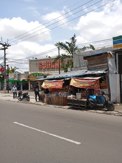 The scene is a street in Katapang, Indonesia, located at the coordinates -6.988 latitude and 107.558 longitude. It is a sunny day, with a clear blue sky and white clouds. In the foreground, there is a black motorcycle parked on the asphalt road. A white line divides the road in half. Behind the motorcycle, there is a small fruit stand on the sidewalk, with a variety of fruits and vegetables displayed for sale. A blue motorized cart sits beside the stand. Three people are visible - two are standing near the fruit stand, and one is standing in the middle of the road near the motorcycle. The stand is shaded by a canopy, and the building behind it has a sign reading "GUDANG" in large red letters. A large green sign near the front of the building reads "MURAH BANGET". The building also has a large white sign with red lettering that reads "GUDANG BARANG". Several power lines run across the sky. There are trees in the background, and a blue and green building can be seen in the distance.