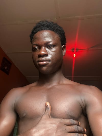 A close-up portrait of a shirtless young Black man with dark, short, curly hair, facing the camera. The man has smooth, dark skin and is slightly glistening. His right hand is placed on his chest. The background consists of an off-white wall with visible seams, and a red light source is visible in the upper part of the image. The location is unspecified but the request contains the city of Mosafejaw, Nigeria. A small rectangular object is on the floor with what looks like a pattern on it.