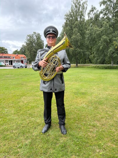 A man wearing a formal grey military-style uniform with a peaked cap and glasses stands in a grassy outdoor area, holding a brass tuba. The man is looking directly at the camera. Behind him, a red-roofed building with white trim and a silver car parked in front of it is visible. The background is filled with green trees under a cloudy sky. The scene appears to be a park or public space in Aneby, Sweden.