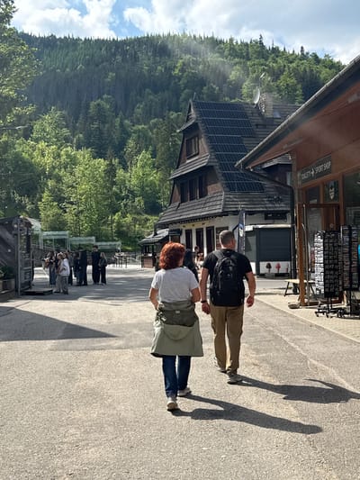 The image depicts an outdoor scene in a vibrant mountain town, likely Zakopane, Poland, under bright sunlight during late morning or early afternoon. The perspective is from slightly behind two individuals walking on a paved pathway, with their long shadows stretching across the ground.
In the immediate foreground, a person wearing a black t-shirt, tan pants, and a black backpack walks beside another person with red hair, a striped shirt, blue jeans, and a light green vest or jacket. Ahead of them, the paved area extends into the midground where several other pedestrians are visible, some strolling and others standing near various establishments.
To the left of the pathway, a row of traditional wooden buildings with intricate architectural details lines the street. One building features several solar panels on its roof and a visible satellite dish. Numerous shopfronts are present, with one displaying racks of souvenirs or postcards. Visible text on signs includes "TOALETY SPORT SHOP" (Toilets Sport Shop) on a brown wooden sign with an arrow, and "RESTAURACJA" (Restaurant) on another sign further down the street. A stand displaying "GOOFY" is also noticeable among the people.
The background is dominated by a dense, verdant forest covering a steep mountainside, extending towards a sky with scattered white clouds. The scene conveys a lively, casual atmosphere typical of a tourist destination nestled in a mountainous landscape.