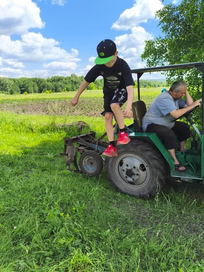 In a rural setting, likely on the outskirts of Ilmino, Russia, a boy wearing a black t-shirt, black shorts with a white pattern, a black and green baseball cap, and red sneakers stands on the back of a green tractor. He is facing away from the camera, with his right arm extended and his fist clenched. An adult male, wearing a grey t-shirt and dark shorts, sits on the tractor's driver's seat, resting his head on his left hand and holding the steering wheel with his right. He is wearing dark sandals. To the left of the tractor, a plow attachment is visible. The ground is covered in green grass, and in the background, there are fields and trees under a blue sky with white clouds.